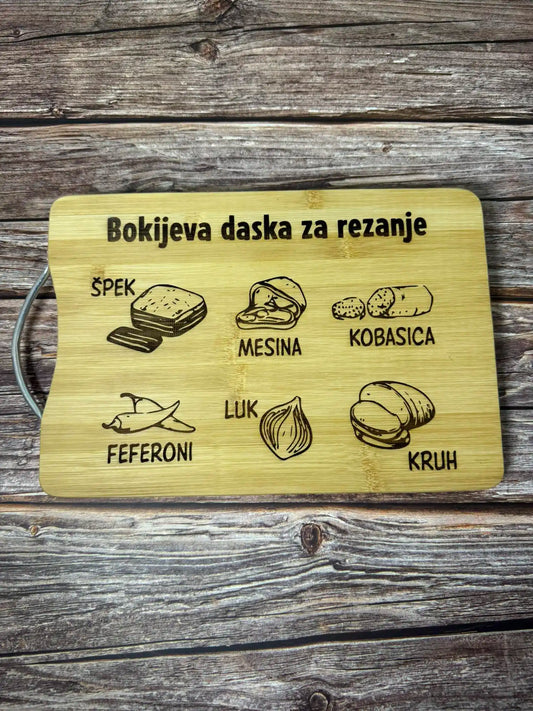 Bokijeva daska za rezanje s humorističnom personaliziranom gravurom koja uključuje nazive namirnica poput špeka, luka, feferona i kruha. Postavljena na rustikalnoj drvenoj podlozi.